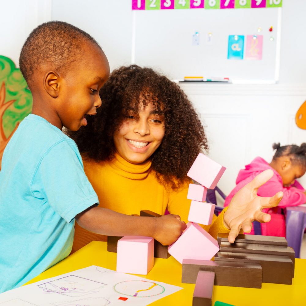 Little black boy play with teacher on building and breaking tower of blocks in nursery class