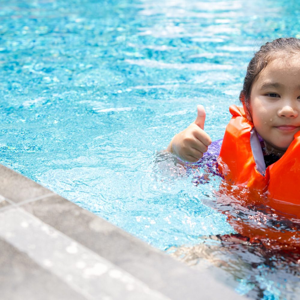 Asian child playing in the pool. Wearing orange life jacket, smiling with thumbs up.