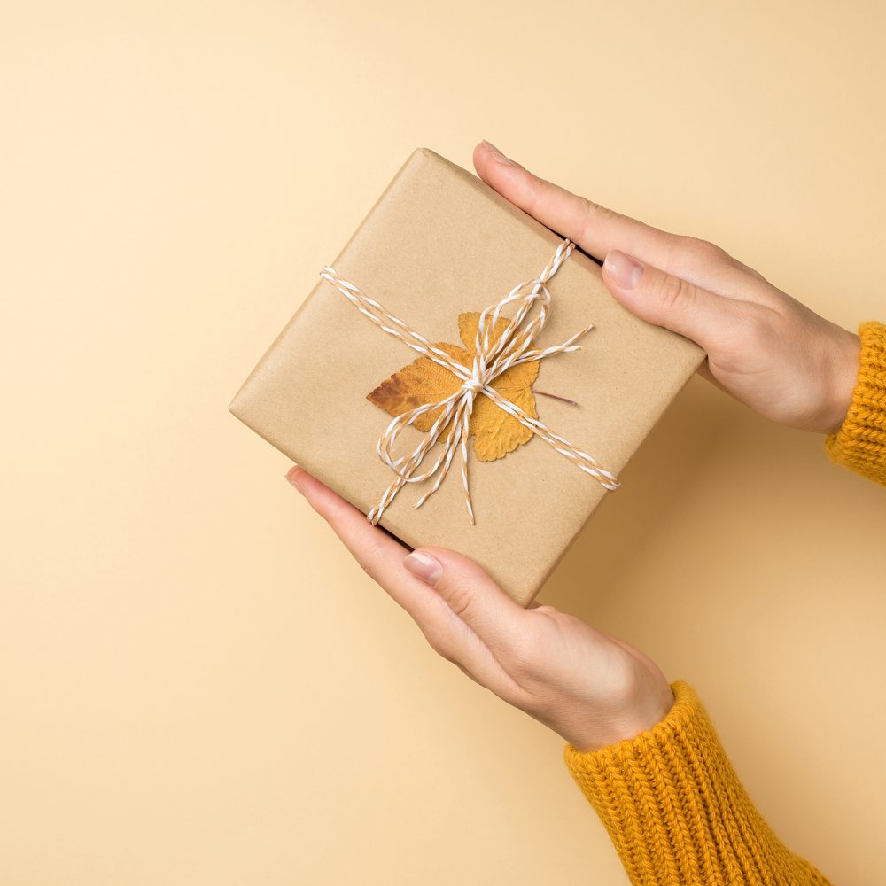 First person top view photo of hands in yellow sweater holding craft paper giftbox with twine bow and yellow autumn leaf on isolated beige background with copyspace