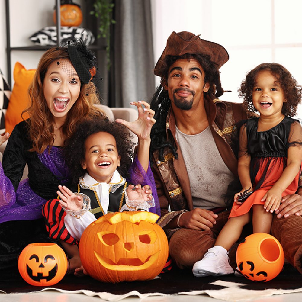 Joyful african american family parents and children in Halloween costumes making scary gesture and smiling at camera while sitting on with jack-o-lantern on floor, celebrating all hallows day