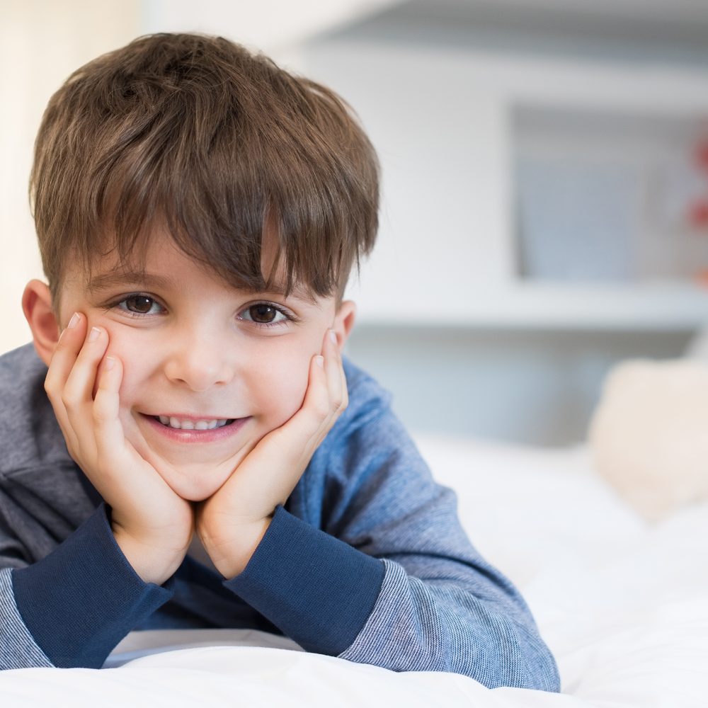 Handsome boy with toothy smile looking at camera. Lovely child lying on front in bed. Happy young cute boy lying on bed.